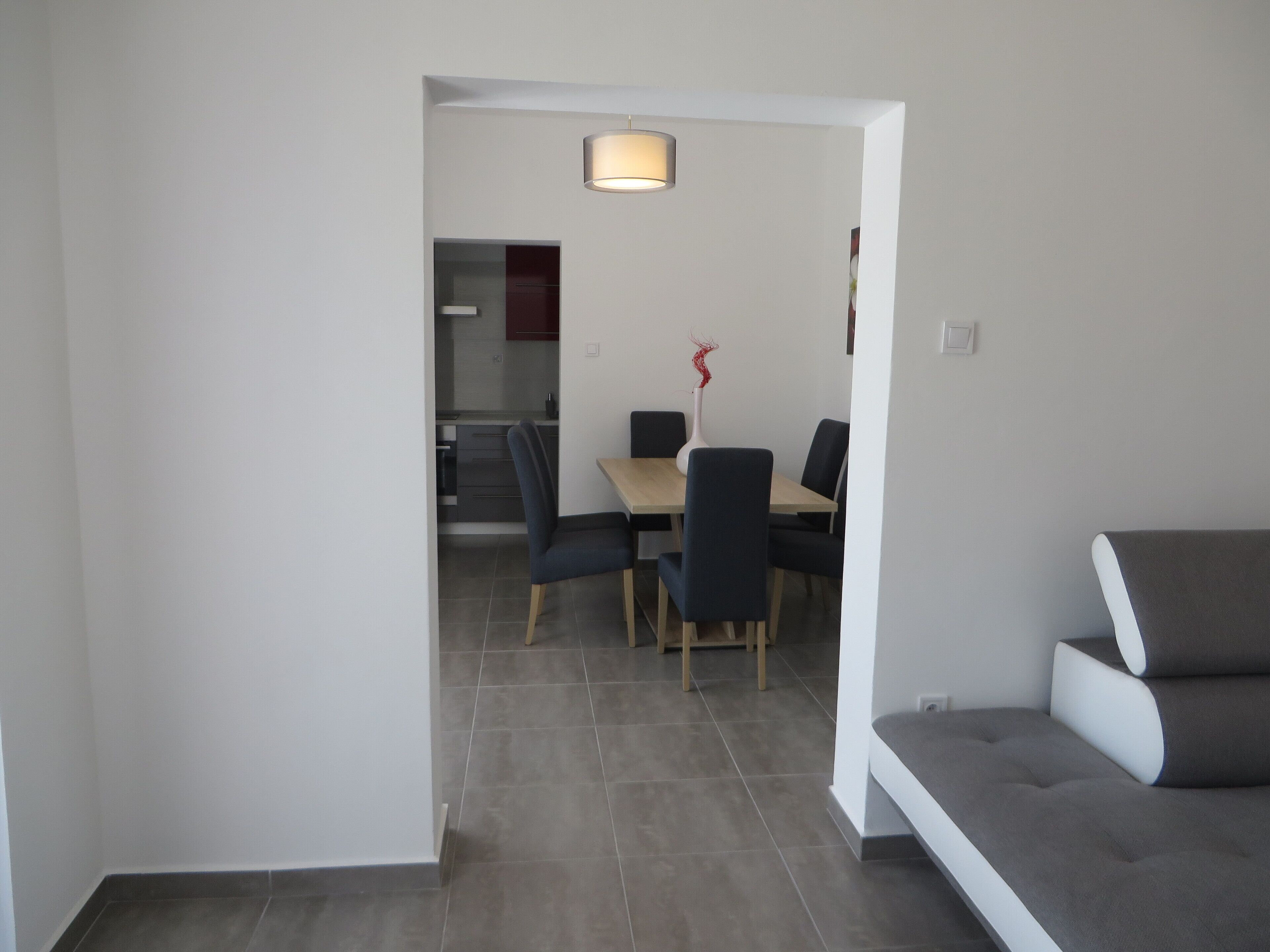 A modern living room with a grey sofa, a dining area with a table and chairs, and a kitchen visible through an open doorway.