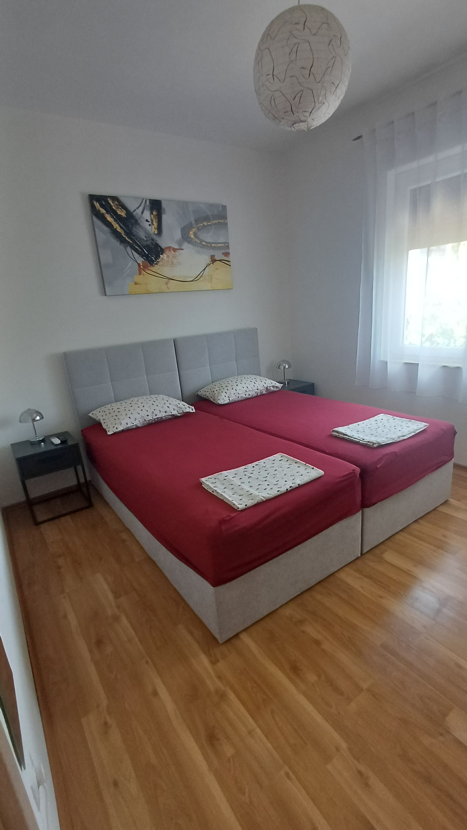 Ein Doppelbett mit roter Bettdecke und zwei Kissen, ein Nachttisch mit Lampe sowie ein Gemälde an der Wand.