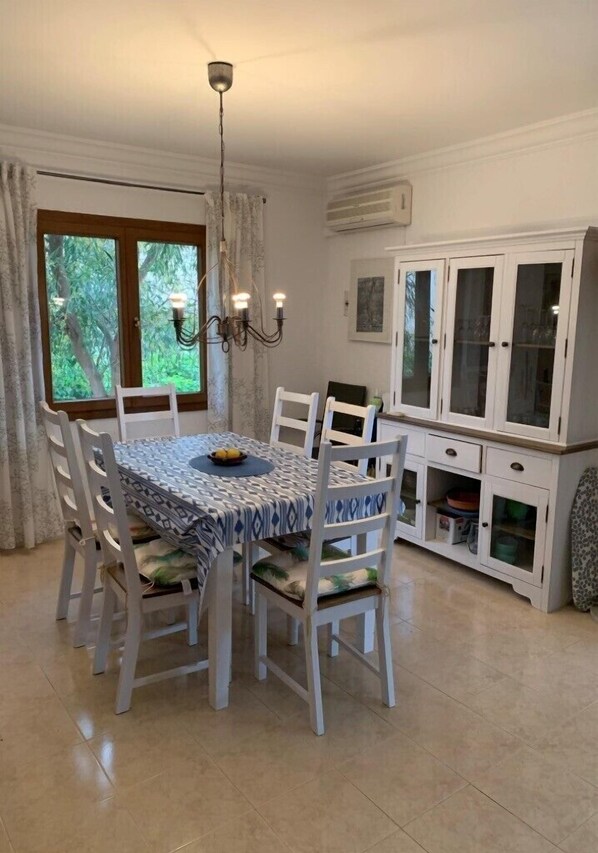 A dining area with a round table, white chairs, and a chandelier. There is a window with curtains, a cabinet with glass doors, and a ceiling fan.