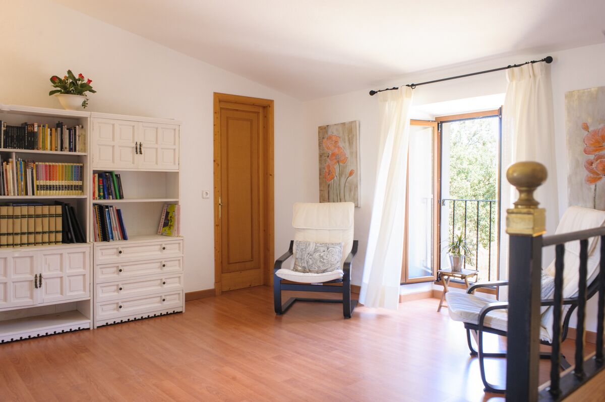 A room with a wooden floor, a white cabinet with books, a chair with a cushion, a door, and a window with curtains.