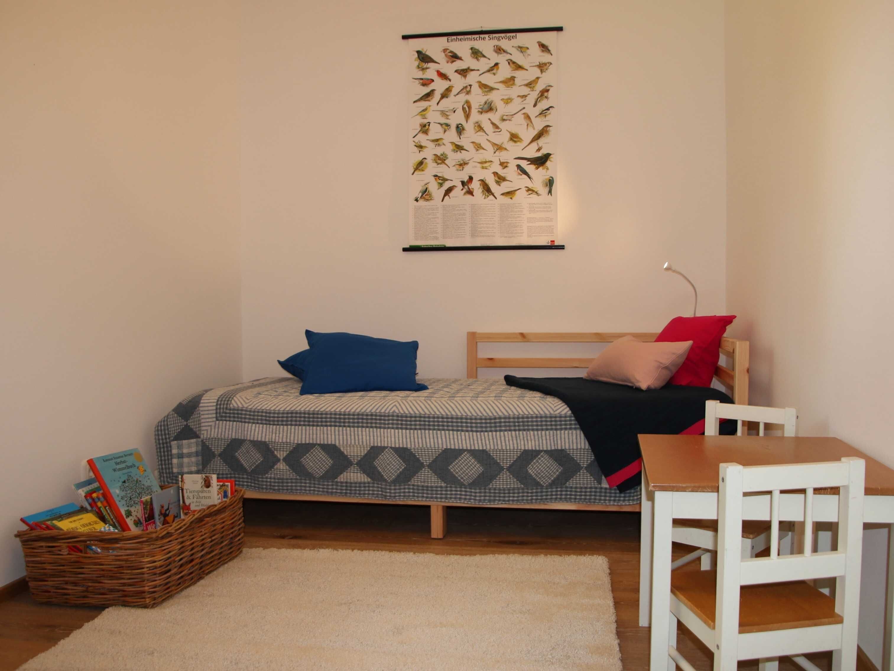 Ein Schlafzimmer mit einem Bett, einem Holz-Kopfteil, einem kleinen Tisch, einem weißen Stuhl, einem Wandposter und einem Korbwäscher mit Büchern.