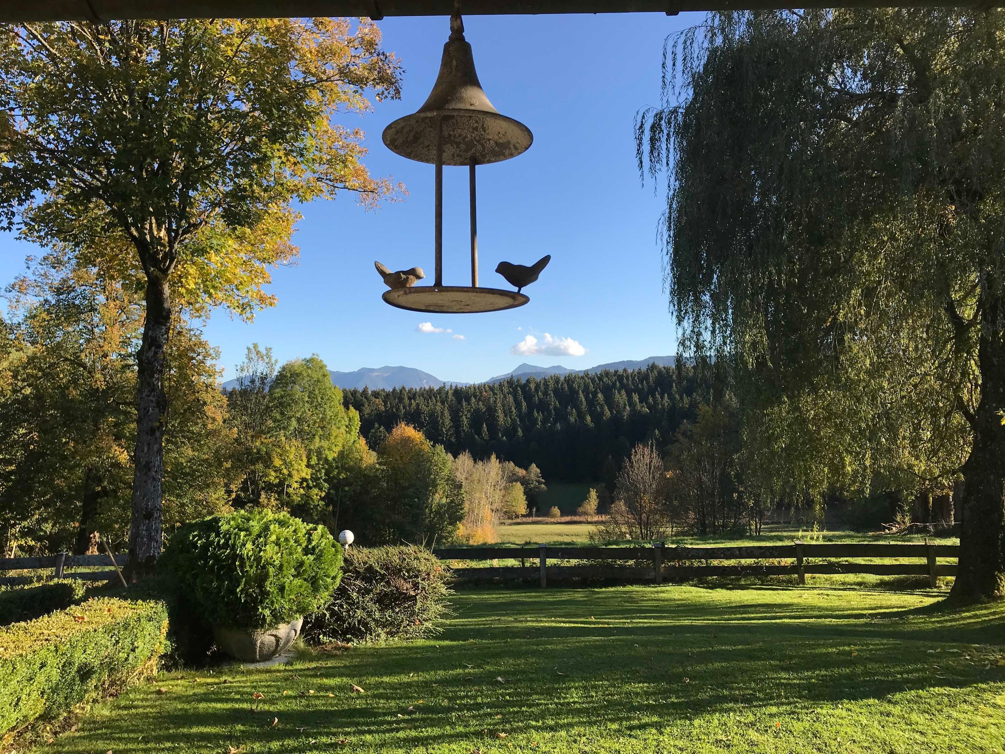 A park with a hanging bell, trees, a wooden fence, and a distant mountain range.
