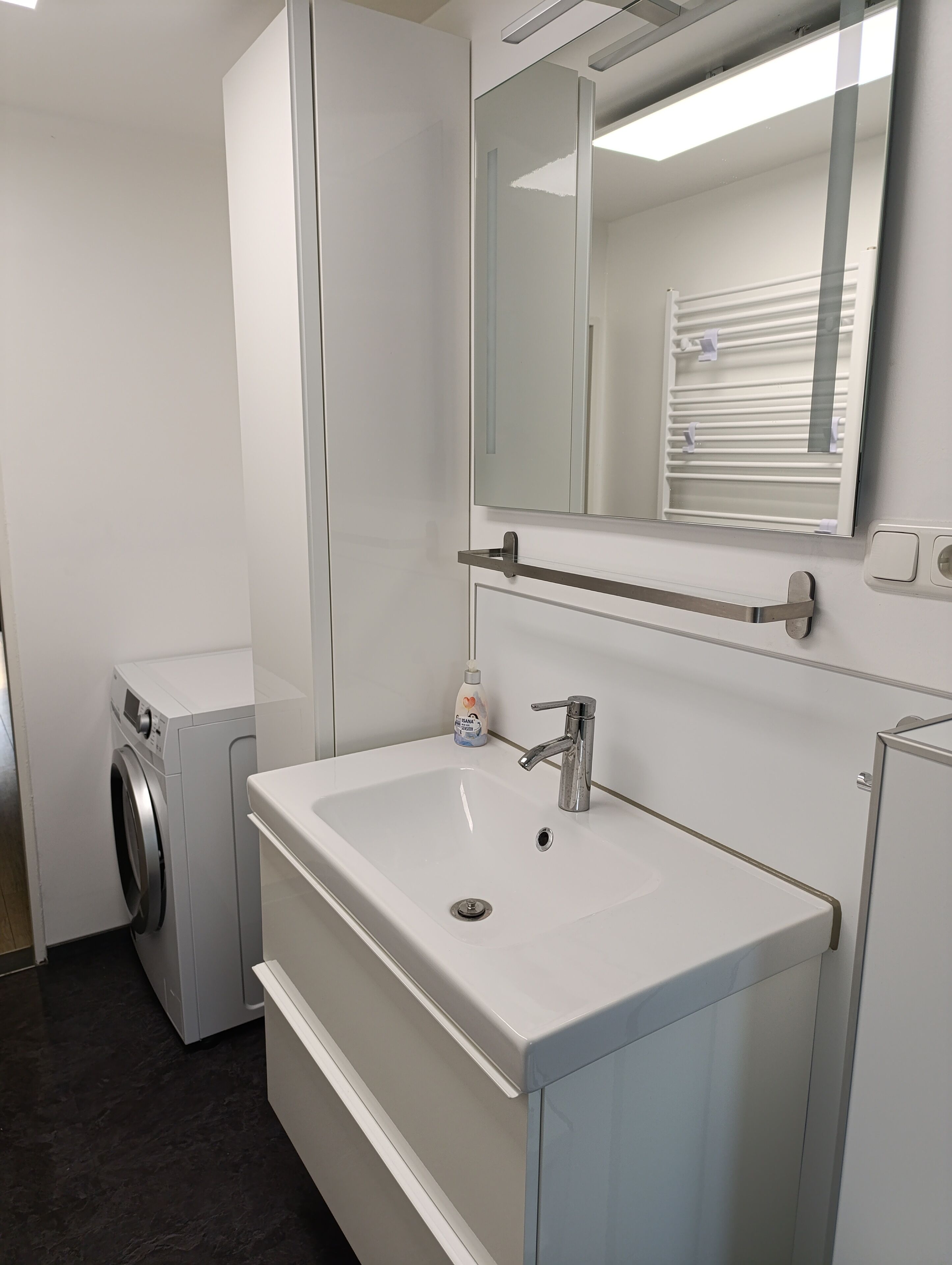 A modern bathroom with a white sink, a mirror, a towel rack, and a washing machine.