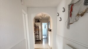 Een hal met een houten vloer, witte muren en een zwarte deurmat bij de ingang.