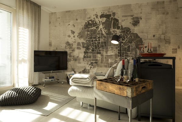 A modern living room with a large TV, a cozy sofa, a wooden coffee table, and a cityscape wall. - FEWO-KARLOTTA New 2-room / 5 min. Ammersee & Zentrum- LUXURY in the heart of Dieessen! (Diessen)