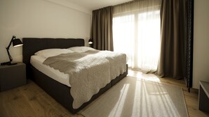 A neatly made bed with a white comforter and a beige blanket, a bedside lamp, a wooden floor, and a large window with curtains. - FEWO-KARLOTTA New 2-room / 5 min. Ammersee & Zentrum- LUXURY in the heart of Dieessen! (Diessen)
