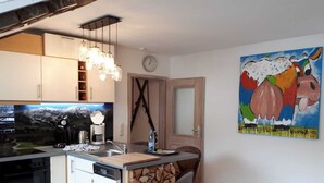 Uma cozinha compacta com fogão embutido, cafeteira e pia. Há uma ilha de madeira com tábua de corte e uma cadeira. Um quadro colorido de uma vaca está na parede.