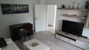Ein modernes Wohnzimmer mit einem weißen Sofa, einem weißen Couchtisch, einem Fernseher auf einem Holzständer, einem Schreibtisch mit Stuhl und einem Gemälde an der Wand.