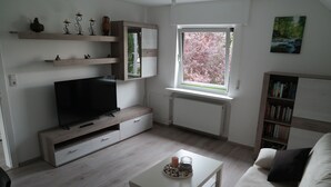 Ein modernes Wohnzimmer mit Flachbildfernseher, Holzböden, einem Fenster mit Blick auf Bäume, einem Bücherregal und einem weißen Sofa.