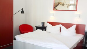 Ein Hotelzimmer mit einem Bett, einem roten Sessel, einem kleinen Tisch mit Lampe sowie einem Bild an der Wand.
