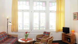 Ein Zimmer mit einer Couch, zwei Sessel, einem Tisch mit Blumenvase, einem Fernseher und einer Lampe.