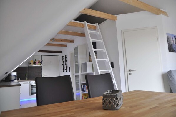 A dining area with a wooden table, black chairs, a white ladder, and a kitchen in the background.