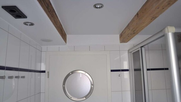 Un bany modern amb rajoles blanques, un mirall rodó, bigues de fusta i una cabina de dutxa de vidre.