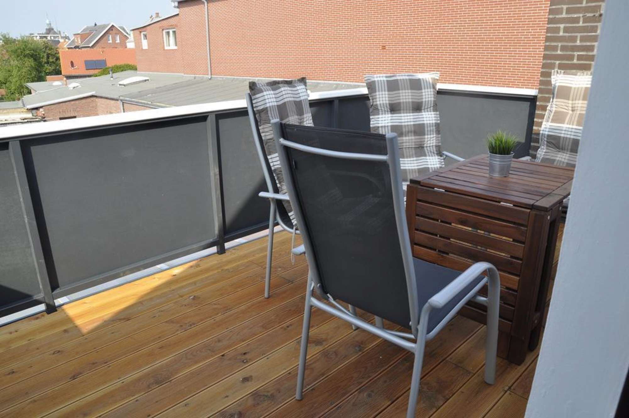 A balcony with two chairs and a small table, a wooden deck, and a brick building in the background.