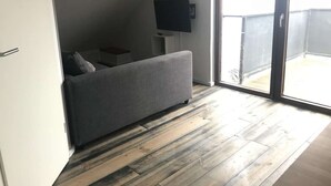 Habitació petita i moderna amb un sofà gris, televisió de pantalla plana i una porta corredissa de vidre que dona a un balcó exterior.