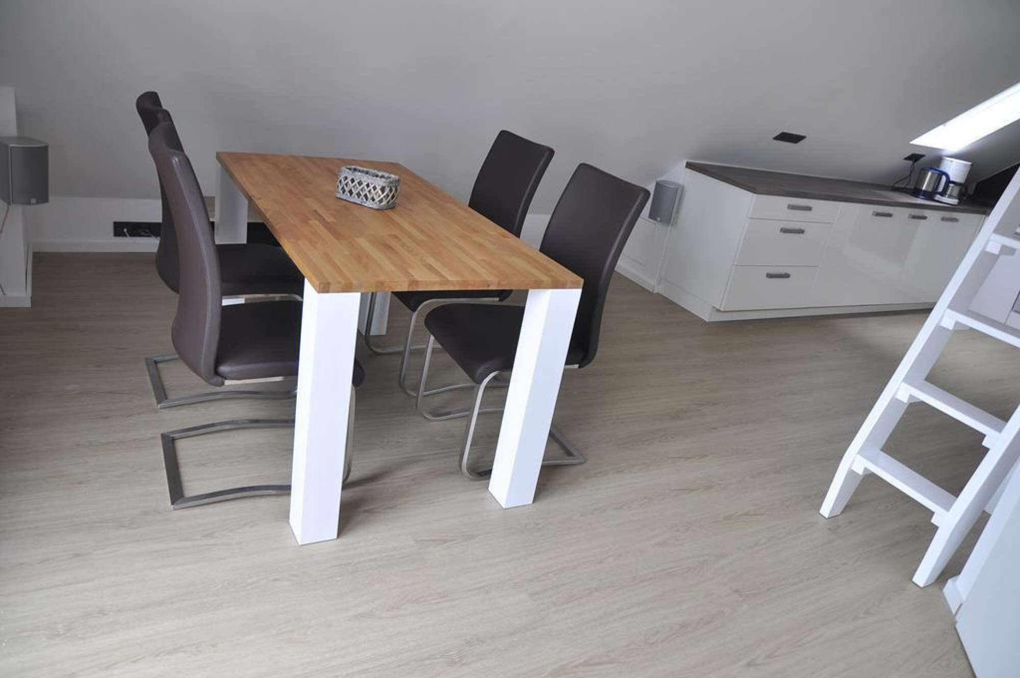 A modern dining area with a wooden table and four chairs, a kitchen area with a white countertop and cabinets, and a white chair with a metal frame.