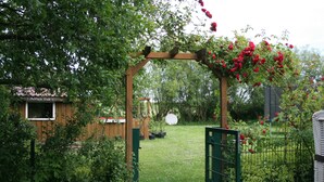 A wooden arbor with red flowers, a green gate, and a wooden structure in the background. - Beautiful vacation apartment with large outdoor terrace near the dike (Simonsberg)