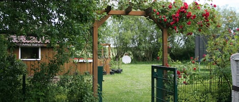 Ein hölzerner Kletterbogen mit roten Blüten, einem grünen Tor und einer Holzkonstruktion im Hintergrund.
