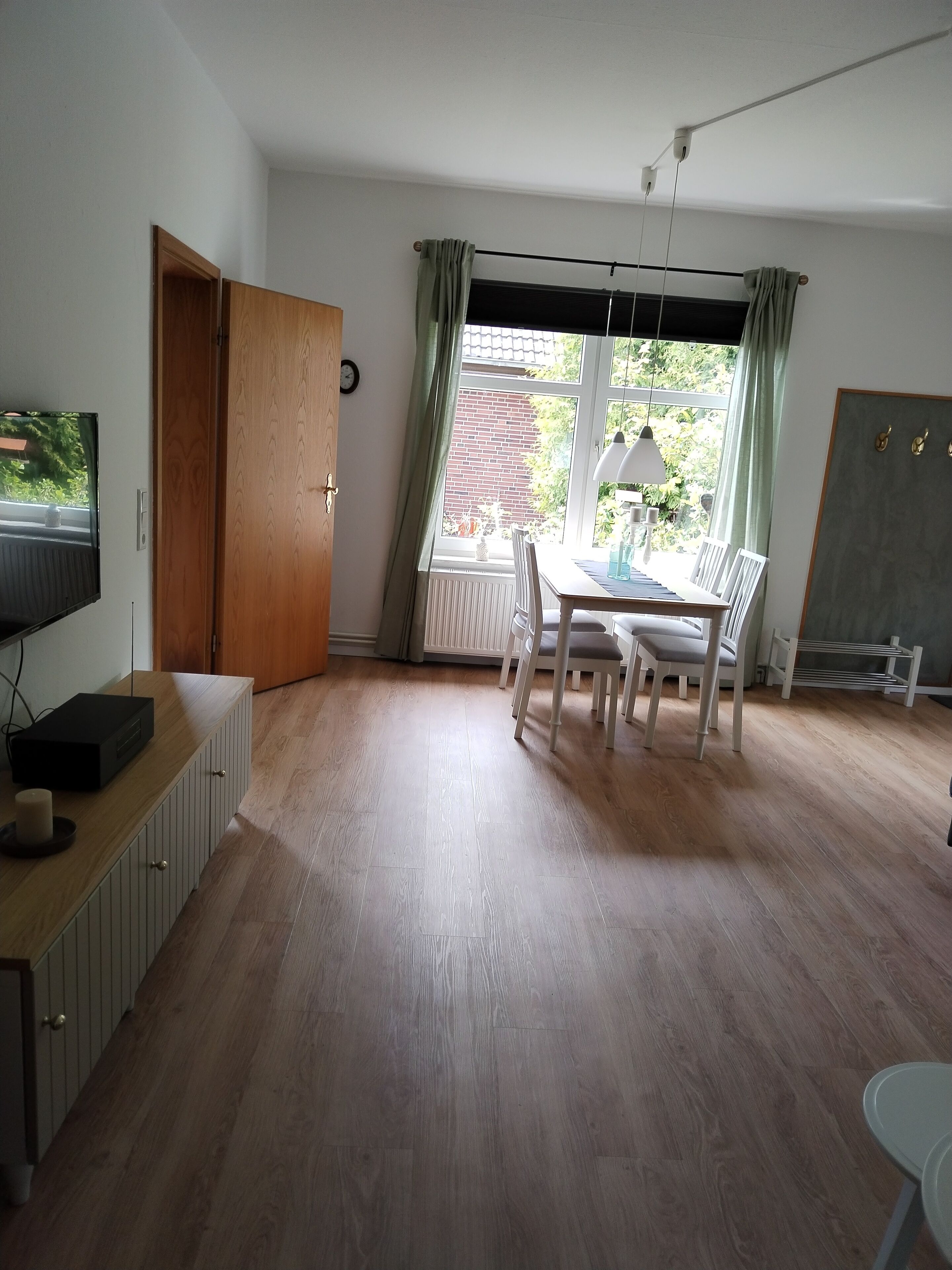 Una habitación con piso de madera, una mesa de comedor con sillas, un televisor sobre un mueble y un ventanal con cortinas.