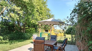 An outdoor patio with a wooden table and chairs, an umbrella, and a view of a yellow building and greenery. - Beautiful vacation apartment with large outdoor terrace near the dike (Simonsberg)