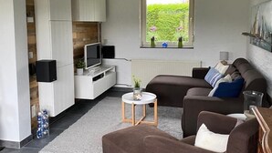 A modern living room with a sofa, ottoman, and a small coffee table. There is a kitchen area with white cabinets and a window with a view of greenery. - Detached holiday home in a quiet location, right on the Grevelingenmeer (Herkingen)