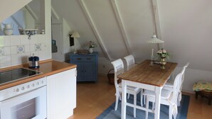 A kitchen with white cabinets, a stove, and a blue cabinet with drawers. There is a dining table with white chairs and a blue rug. - Landhaus Darss - in love with nature (Wieck a. Darß)