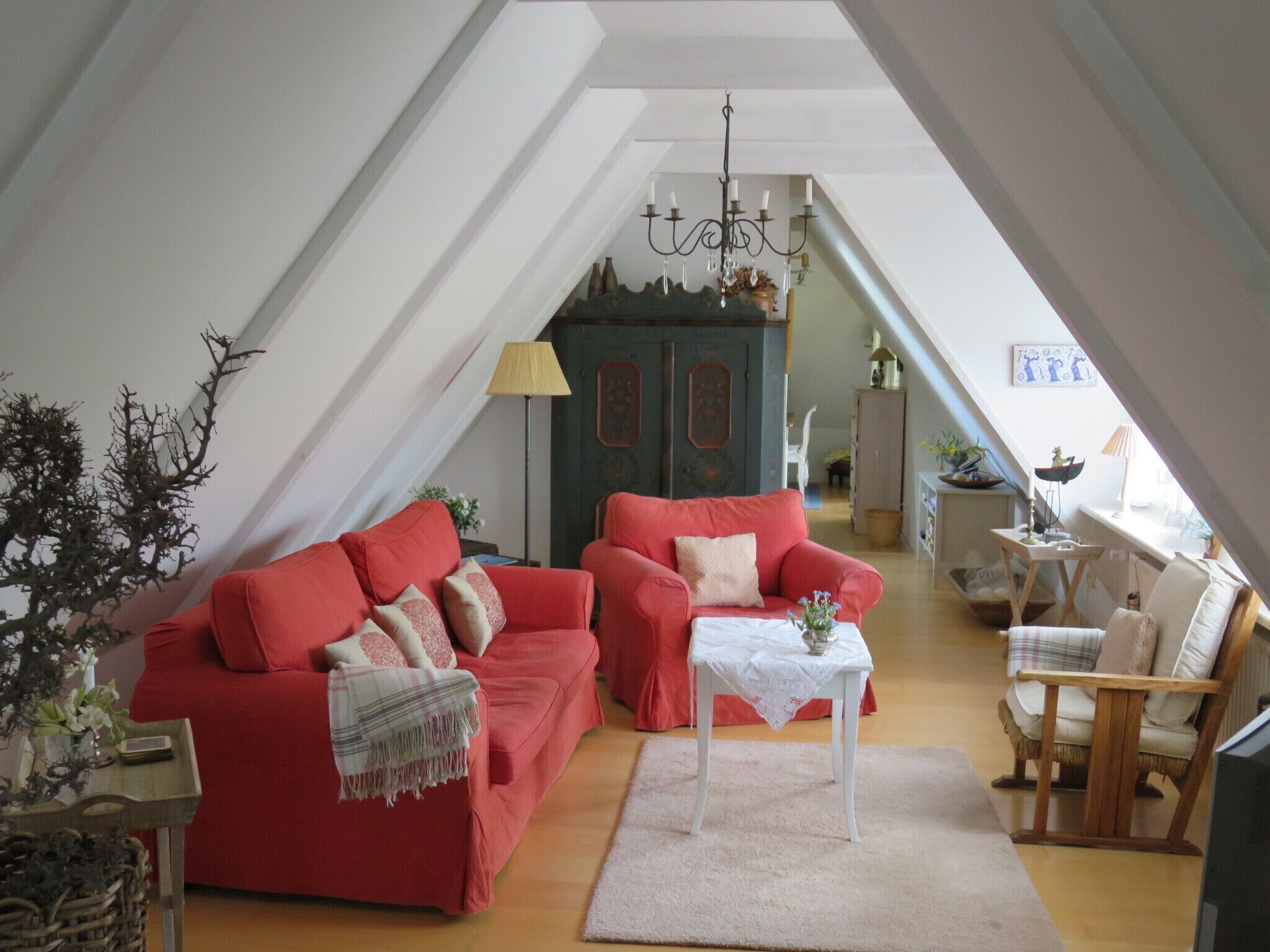 A cozy loft-style living room with a red sofa, a small white table, and a wooden chair. A chandelier hangs from the ceiling, and there is a staircase leading to an upper level.
