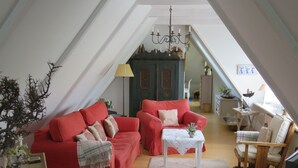 A cozy loft-style living room with a red sofa, a small white table, and a wooden chair. A chandelier hangs from the ceiling, and there is a staircase leading to an upper level. - Landhaus Darss - in love with nature (Wieck a. Darß)