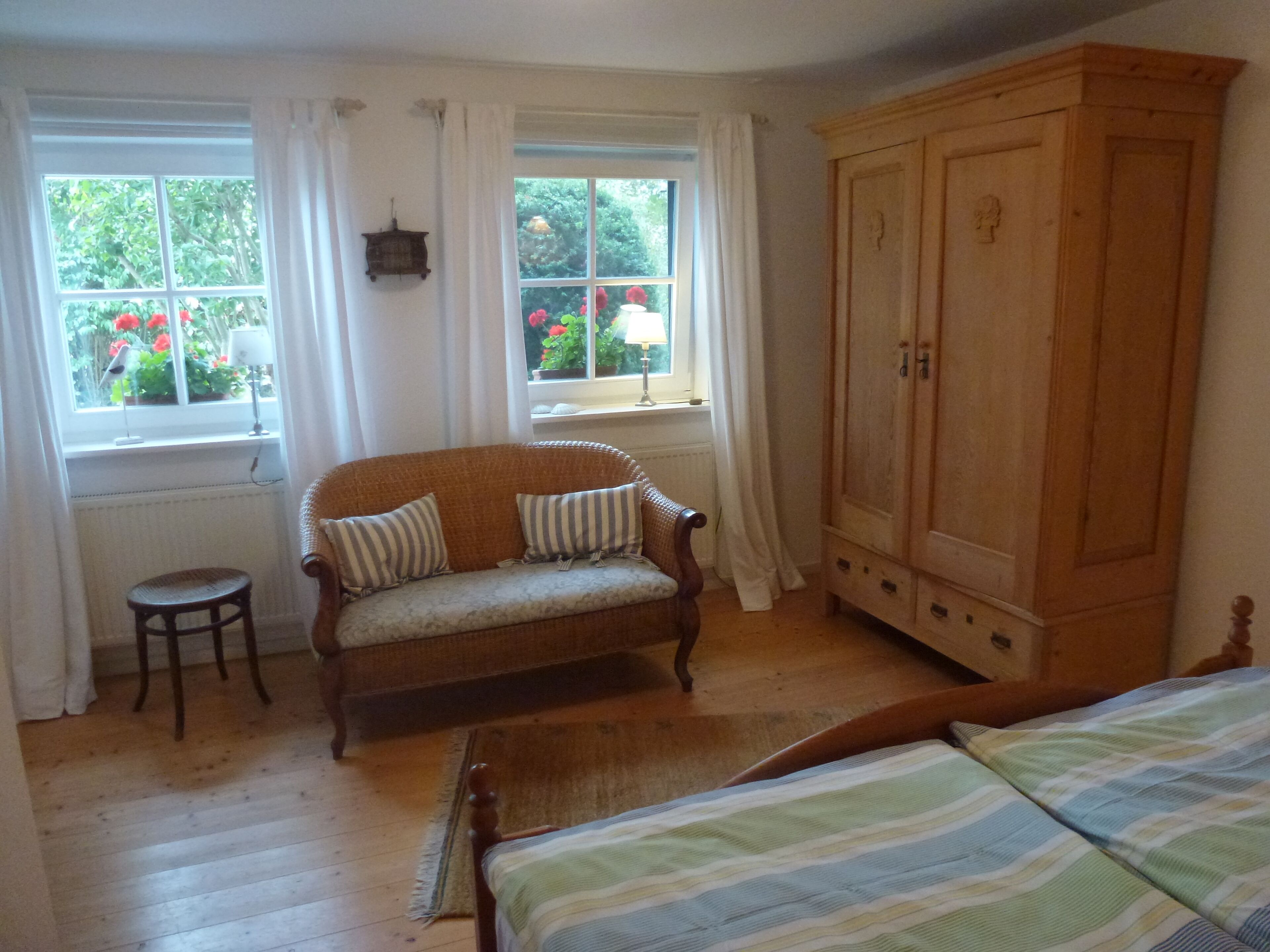 A room with a wooden floor, a wooden bed, a wooden wardrobe, a wicker sofa with striped cushions, a small wooden stool, and two windows with white curtains.