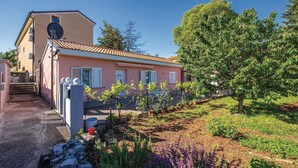 Una casa a due piani con facciata rosa, tetto in tegole e un giardino con diverse piante e un albero.