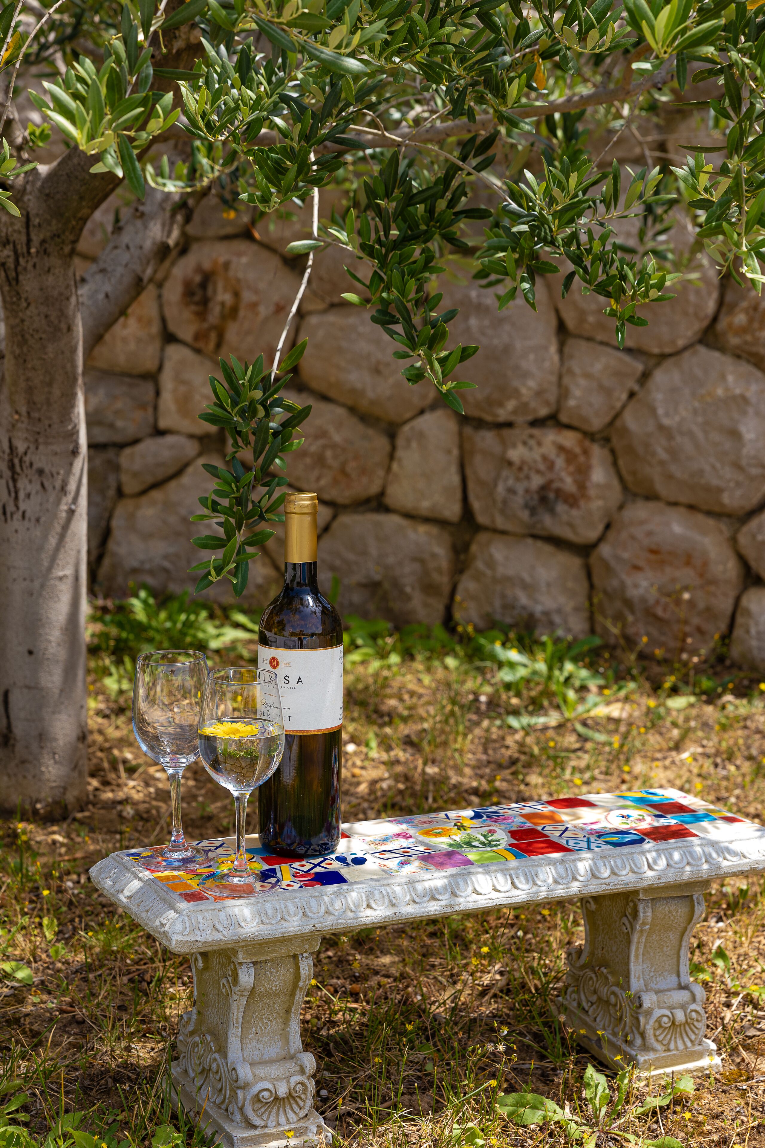 A stone wall, a bottle of wine, and two glasses on a decorative table.