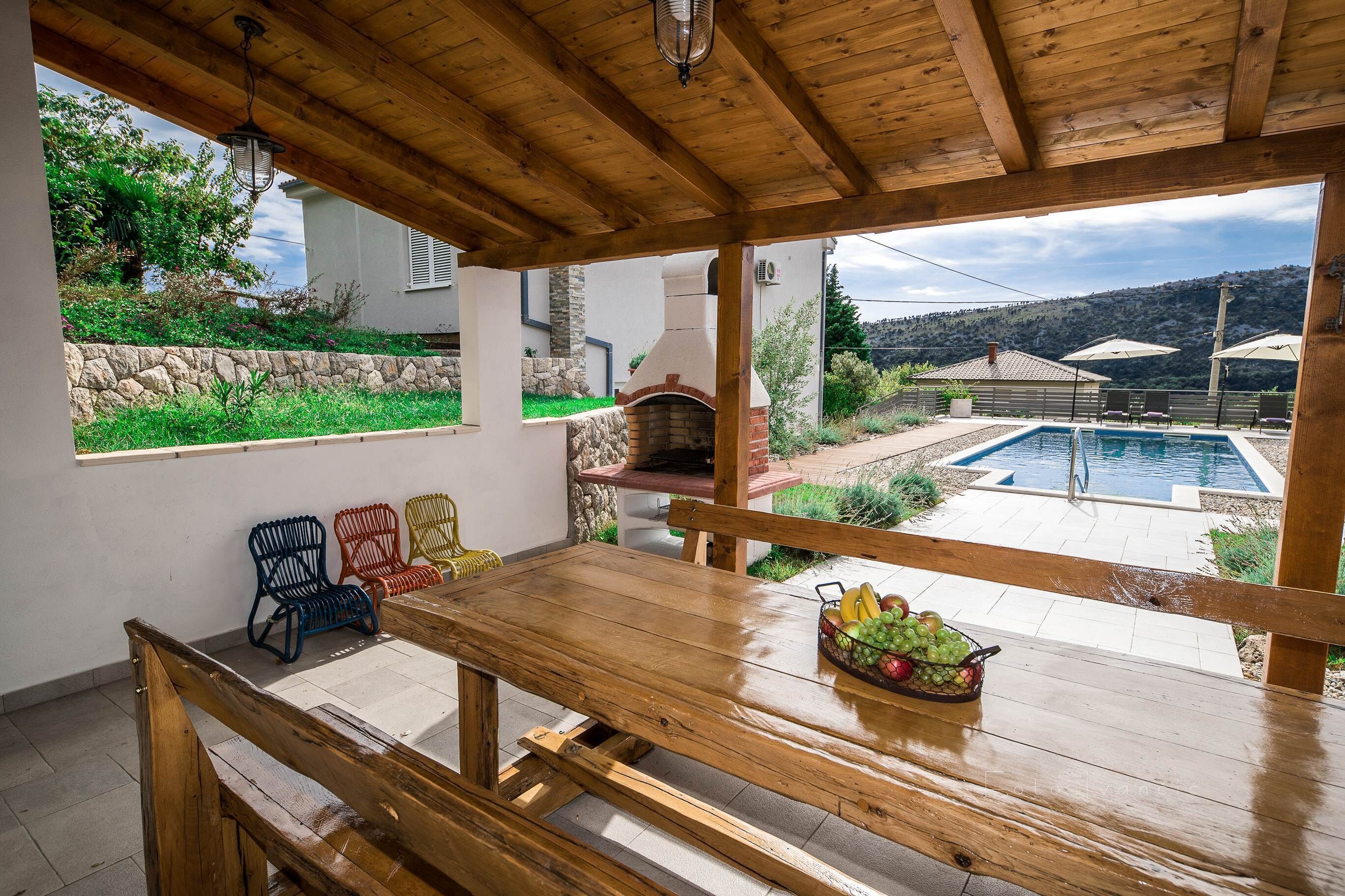 A wooden dining table with a fruit basket, a stone wall, and a view of a swimming pool and hills.