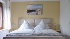 Une chambre à coucher avec un lit, deux tables de chevet, une lampe, un tableau représentant une plage et une fenêtre avec des rideaux.