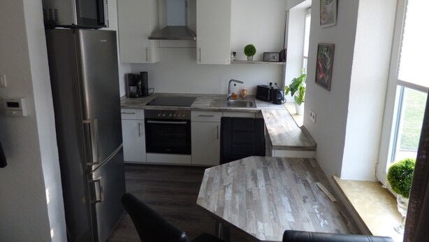 A modern kitchen with white cabinets, a stainless steel refrigerator, a built-in oven, a microwave, and a coffee maker. There is a wooden dining table with chairs. - Apartment on the North Sea (269) (Wilhelmshaven)