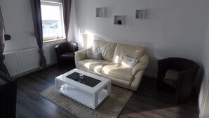 A living room with a beige sofa, a white coffee table, a dark armchair, and a small side table. There is a window with curtains, a radiator, and a lamp. - Apartment on the North Sea (269) (Wilhelmshaven)