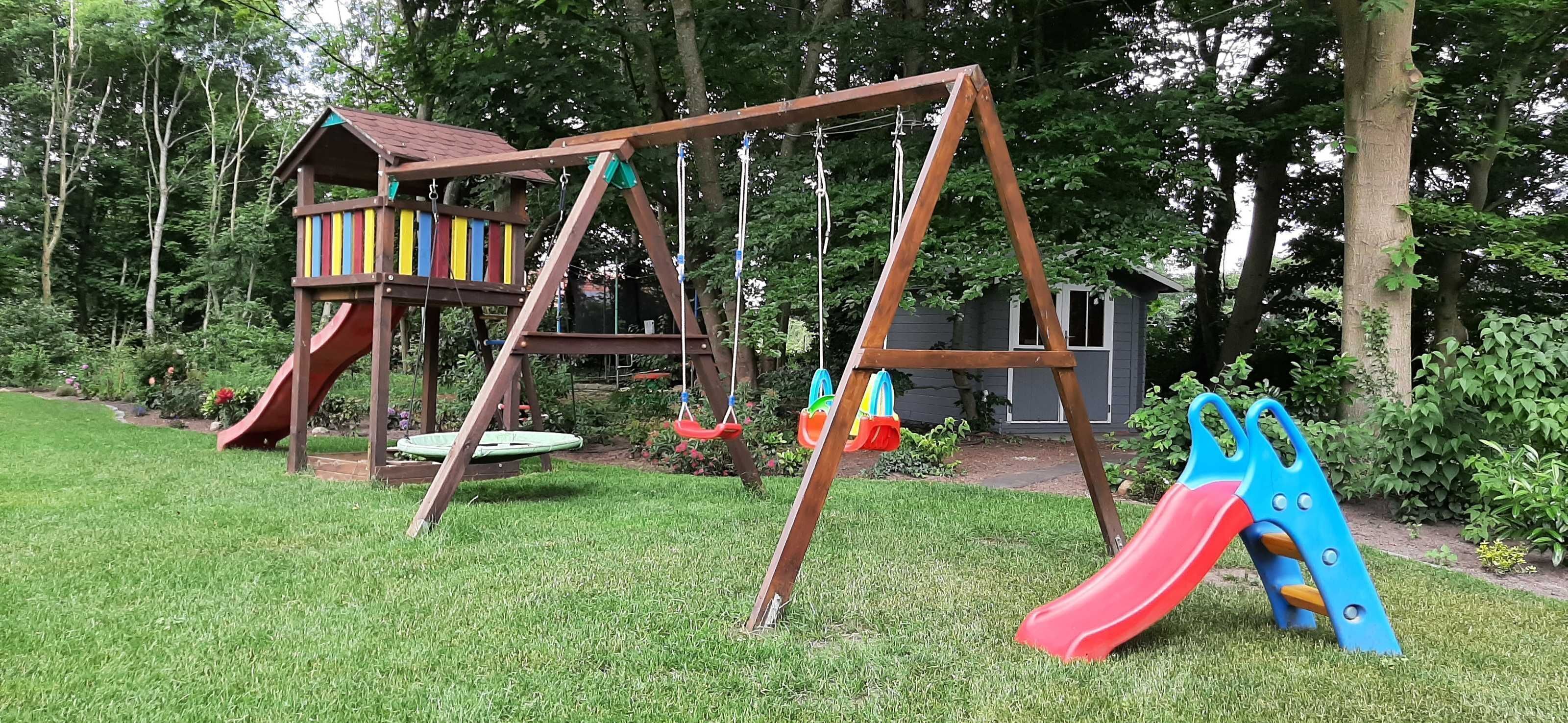 Een houten schommelset met een glijbaan, een klein boomhuis en een blauwe klimconstructie in een gazon met bomen op de achtergrond.