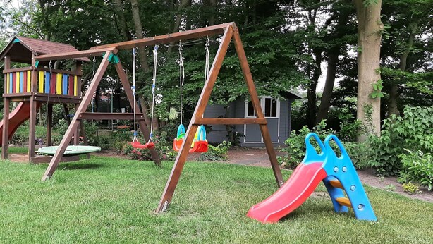 Um baloiço de madeira com escorrega, uma casinha de árvore pequena e uma estrutura azul para escalar num jardim relvado com árvores ao fundo.