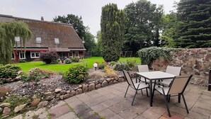 Um pátio com mesa e cadeiras, um muro de pedra e um jardim com diversas plantas e gramado.