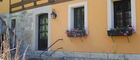 A building with a yellow facade, stone base, and timber framing. There are two windows with black shutters, one with a flower box.