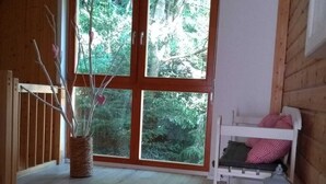 Ein Zimmer mit großem Fenster, Holzwänden, einem weißen Bett mit rot-weißen Kissen, einem kleinen weißen Nachttisch und einem Korbwannenkorb mit Ästen.