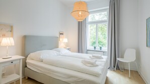 Ein modernes Schlafzimmer mit Bett, Nachttischlampen, einem Fenster mit Vorhängen, einem Sessel und einer Pendelleuchte.