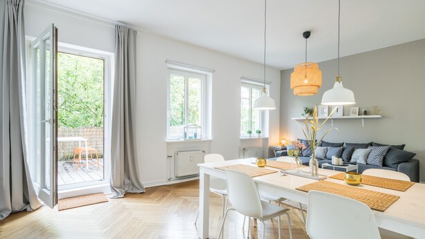 Ein helles, modernes Wohnzimmer mit einer großen Schiebetür zum Balkon, einem weißen Esstisch mit Stühlen, einem grauen Sofa und einer weißen Decke.