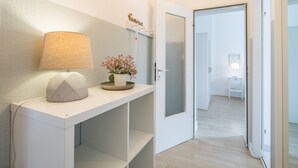 Ein Flur mit einem weißen Konsolentisch, einer Lampe und einer kleinen Pflanze. Eine Tür mit Sandstrahlglas führt in einen weiteren Raum.