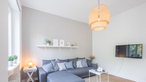 Ein modernes Wohnzimmer mit einem grauen Ecksofa, einem weißen Beistelltisch mit Lampe, einem an der Wand montierten Fernseher und einem Regal über der Couch.