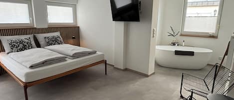 A modern bedroom with a bed, a flat-screen TV, a bathroom with a bathtub, and a desk with a chair.