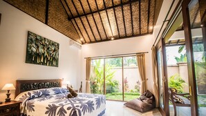 A bedroom with a large bed, a nightstand, a window with curtains, and a view of the garden. - Sujaya Villas (Ubud)