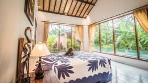 A bedroom with a large bed, a lamp, and a view of a lush garden and pool. - Sujaya Villas (Ubud)