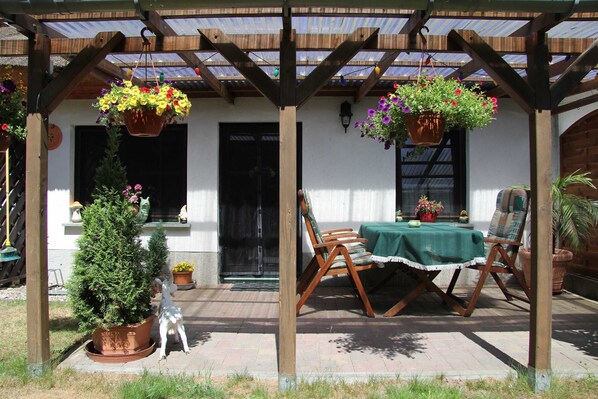 A covered patio with a table and chairs, potted plants, and hanging flower baskets. - Living under the pipe roof (Zempin)
