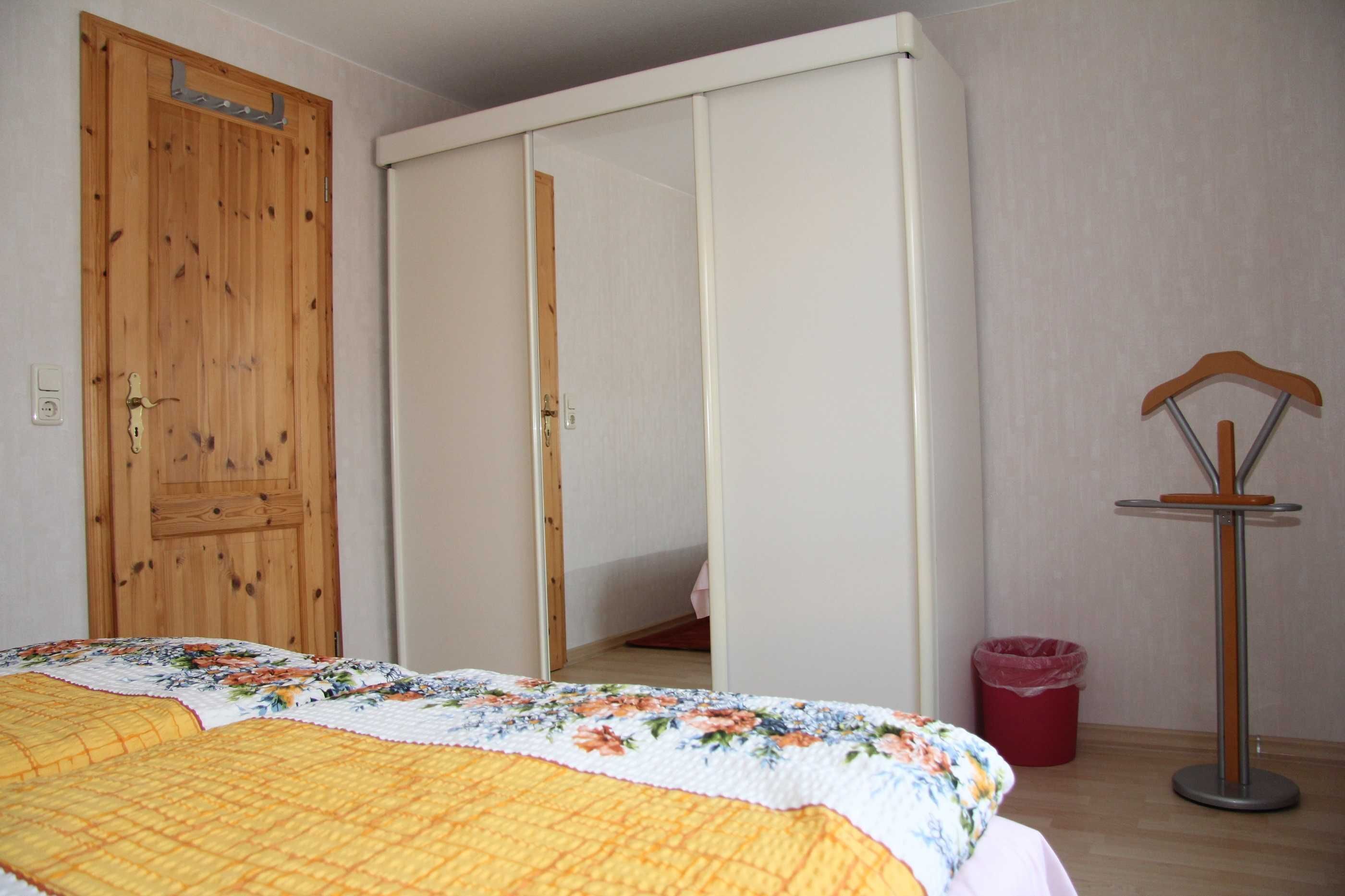 Une chambre avec une porte en bois, une armoire, un lit recouvert d’une couette à motifs floraux et une poubelle rouge.