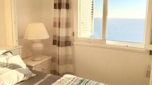 A bedroom with a bed, a nightstand, a lamp, a window with shutters, and a view of the sea. - Jandia Mar 2 (Morro Jable)
